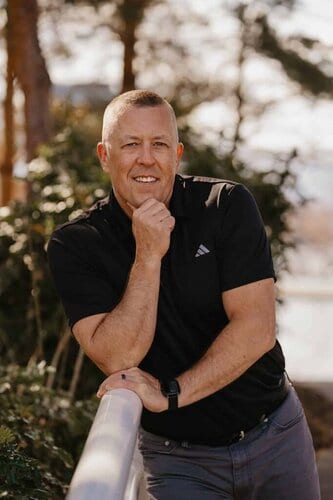 A smiling man standing outdoors near a waterfront, enjoying a peaceful moment in nature, representing mental health and well-being in counselling.