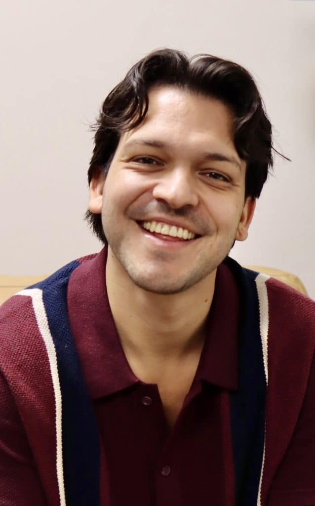 Smiling male therapist with dark hair in a maroon and navy sweater, sitting in a counselling office.