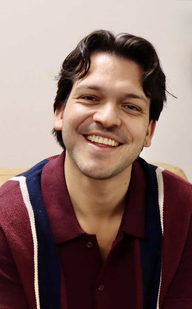 Smiling male therapist with dark hair in a maroon and navy sweater, sitting in a counselling office.